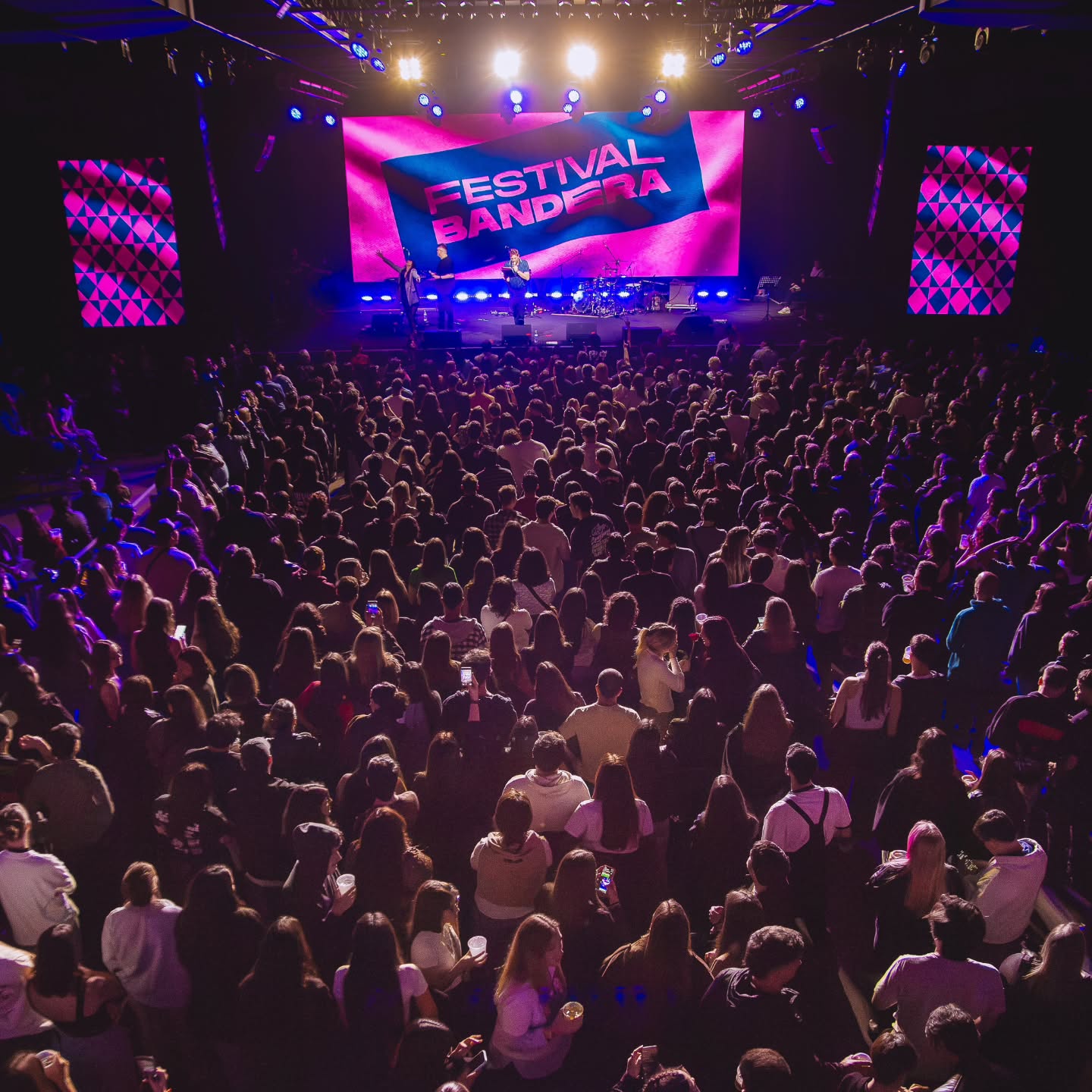 Una toma aérea de un concierto en un predio cerrado o semi-cubierto, repleto de gente de espaldas de cara a un escenario iluminado. En el fondo, una gran pantalla LED de color magenta brillante muestra el texto "FESTIVAL BANDERA" en letras oscuras, mientras se distinguen músicos actuando en la tarima bajo luces cálidas de reflectores.