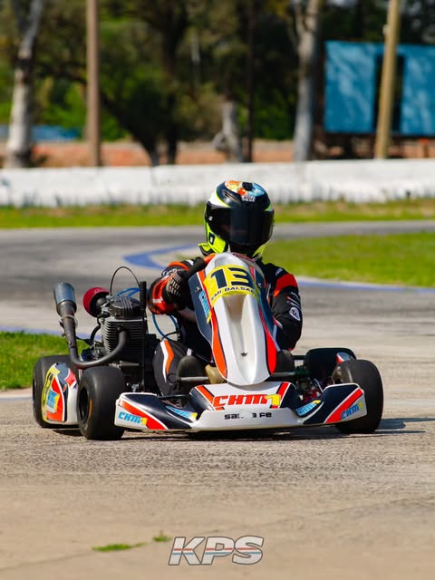 Fotografía en primer plano de un piloto compitiendo en una pista de karting al aire libre. El conductor viste un traje deportivo negro con detalles naranjas y blancos, guantes oscuros y un casco con un diseño colorido en la parte superior. El kart lleva el número 13 en el frente, calcomanías comerciales y la inscripción Santa Fe en el paragolpes delantero. En el borde inferior de la imagen se observa de forma centrada el logotipo de la categoría, que dice KPS.